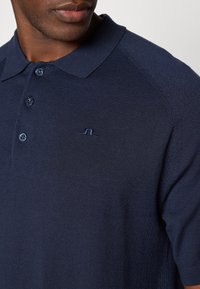 Navy blå polo-skjorte i teksturert strikkemateriale med klassisk krage, som har tre knapper og en liten brodert logo på brystet.