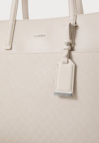 Calvin Klein tote bag in light beige, faux leather with embossed geometric pattern, featuring sturdy handles and a tag with metal accent.