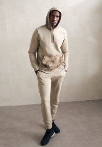 Sudadera beige con bolsillo frontal de patrón camuflaje, combinada con pantalones beige a juego. Usada con zapatillas deportivas negras. Ajuste y diseño casual.