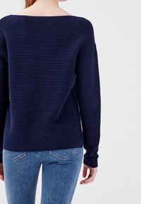 Tmavě modrý žebrovaný svetr se širokým výstřihem a dlouhými rukávy, doplněný světle modrými džínami s jemným vzorem na kapsách.