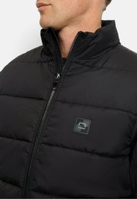 Schwarze Pufferjacke mit hohem Kragen, ausgestattet mit einem Frontreißverschluss und einem kleinen Logopatch auf der linken Brust. Glatte, gepolsterte Textur.