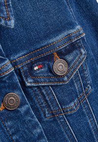 Giubbotto di jeans in tessuto blu scuro, con cuciture arancioni, due tasche sul petto con bottoni metallici e un piccolo dettaglio del logo ricamato.