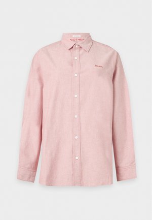 Persona joven que lleva una camisa rosa suelta abotonada sobre una camiseta negra, vaqueros grises de pierna ancha y zapatos negros con punta cuadrada, de pie frente a un fondo sencillo.
