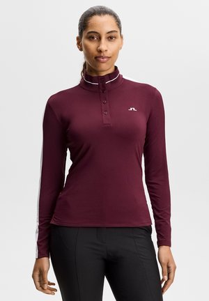 Donna che indossa una camicia bordeaux a maniche lunghe con colletto abbottonato e logo bianco sul petto, abbinata a pantaloni neri, in piedi davanti a uno sfondo semplice.