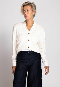 Cremfarbener, verkürzter Cardigan mit V-Ausschnitt, fünf goldenen Knöpfen und gerippten Bündchen. Kombiniert mit dunkelblauen Cargo-Hosen aus Denim mit Seitentaschen.