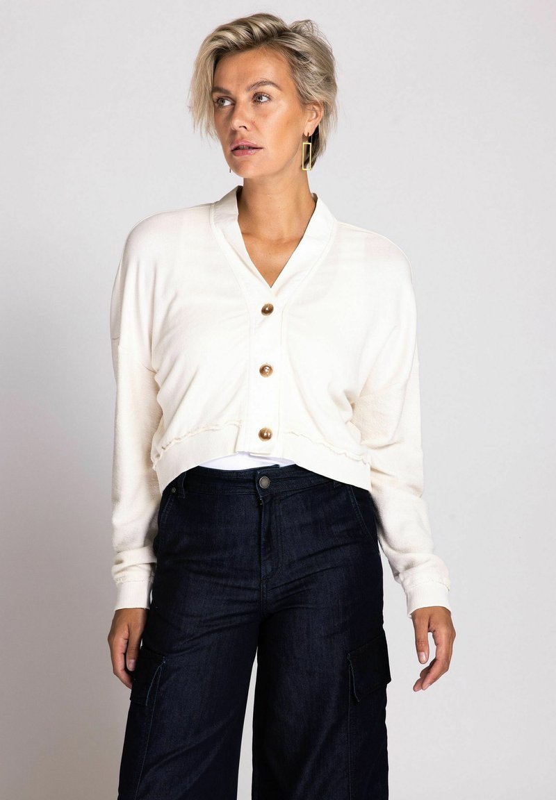 Cremfarbener, verkürzter Cardigan mit V-Ausschnitt, fünf goldenen Knöpfen und gerippten Bündchen. Kombiniert mit dunkelblauen Cargo-Hosen aus Denim mit Seitentaschen.