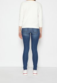 Maglione bianco strutturato con maniche lunghe, abbinato a jeans skinny blu e sneakers bianche con accenti rossi e rosa.