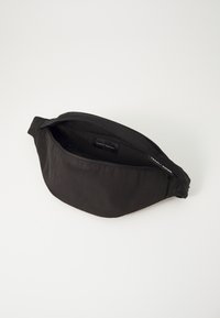 Svart stoff bærenett med en buet form, glidelåslukking og innvendig etikett. Har en jevn tekstur og justerbar stropp for passform.