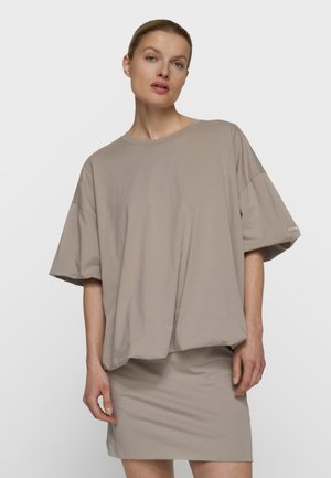 Donna che indossa una maglietta ampia oversize color taupe a maniche corte abbinata a una gonna aderente coordinata, in piedi davanti a uno sfondo chiaro e uniforme.