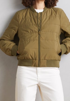 Persoon draagt een olijfgroene doorgestikte bomberjack met ritssluiting aan de voorkant en geribbelde boorden, gecombineerd met witte broek, handen in de zakken van het jack.