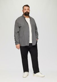 Chemise à carreaux grise et noire sur un t-shirt blanc, associée à un pantalon noir et des baskets blanches, se tenant sur un fond uni.