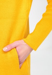 Žlutý pletený svetr s texturovaným povrchem. Jsou viditelné dlouhé rukávy a boční kapsa, které ukazují detail ruky.