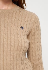 Pull beige en maille torsadée avec manches longues et petit logo brodé bleu marine sur la poitrine sur fond uni.