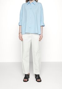 Camicia azzurro chiaro con colletto e maniche arrotolate, abbinata a pantaloni bianchi larghi e sandali con zeppa neri aperti davanti.