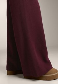 Pantaloni largi din tricot de culoare burgund închis, cu textură ribbed, asortați cu o încălțăminte cu platformă de culoare maron deschis.