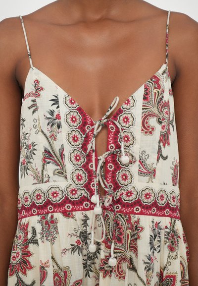 Robe à motifs floraux avec des bretelles spaghetti, présentée avec un décolleté en V profond et une fermeture décorative à nouer avec des pompons. Couleurs principalement crème et rouge.