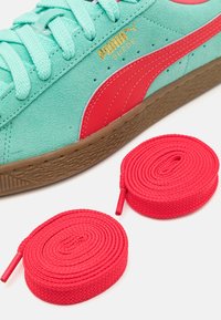 Gros plan d'une basket Puma en daim turquoise avec une bande rouge et un logo doré, à côté de deux lacets rouges enroulés sur une surface blanche.