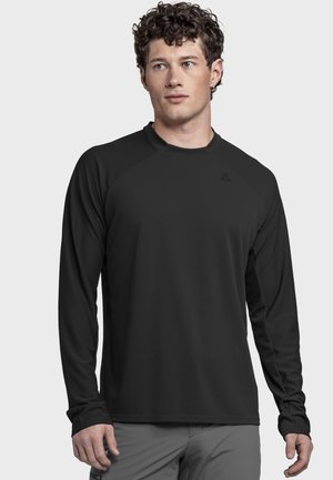 Junger Mann mit lockigem Haar, der ein schwarzes langärmliges Sportshirt und graue Hose trägt, vor einem schlichten grauen Hintergrund stehend.