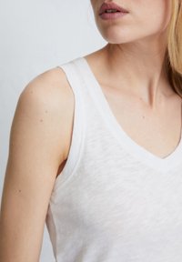 Débardeur blanc fabriqué en tissu léger et légèrement texturé. Il dispose d'un col en V profond et de bretelles étroites, mettant en valeur un design simple.