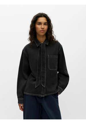 Femme aux cheveux bouclés portant une chemise noire à manches longues boutonnée avec poche poitrine et pantalon foncé, debout devant un fond clair uni.