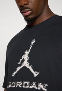 Sort bomulds-T-shirt med et hvidt Jordan-logo grafik med stiliserede detaljer. Rund hals og korte ærmer. Teksten nedenfor lyder "JORDAN."