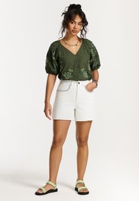 Blouse à manches bouffantes vert olive avec broderies florales, associée à des shorts en denim blancs et des sandales vert clair. Look naturel et décontracté.