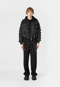 Chaqueta bomber negra con detalles de cintas en capas, combinada con una sudadera con capucha negra y pantalones, que presentan un acento de cadena y calzado texturizado.