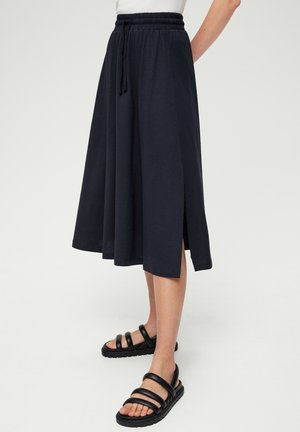 Kvinde iført en sort elastisk midi-nederdel med sidespids, kombineret med sorte flade sandaler med stropper og en hvid top.