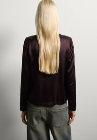 Persona con cabello rubio largo vistiendo una camiseta oscura de manga larga y jeans desgastados de color azul, de pie frente a una pared gris claro lisa.