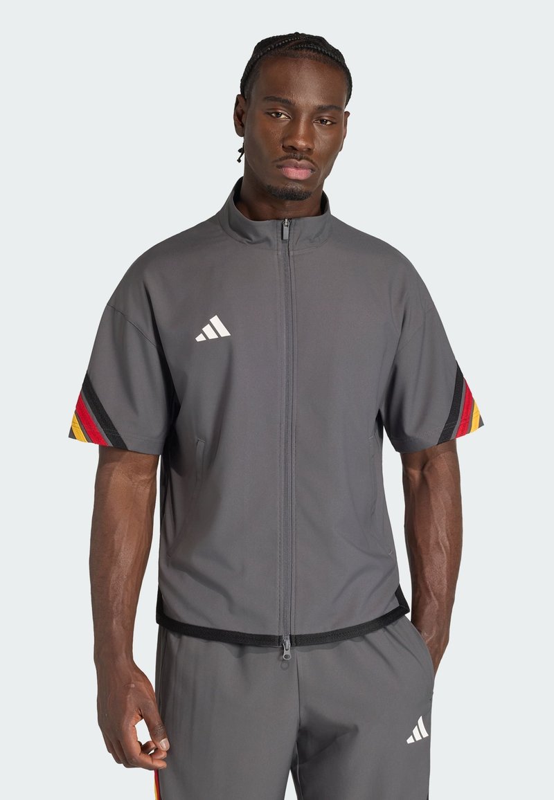 Mann mit geflochtenem Haar, der eine graue kurzärmelige Jacke mit Reißverschluss und Hosen mit Adidas-Logos sowie schwarzen, roten und gelben Streifen an den Ärmeln in neutraler Pose trägt.