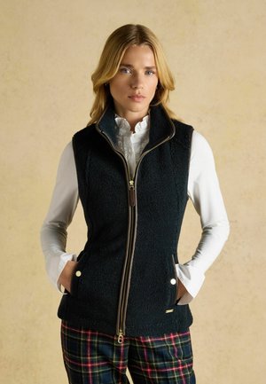 Sort fleece vest med høj krave, lynlås foran og sidelommer. Brun kant med detaljer og en tekstureret stof. Kombineret med ternede bukser.