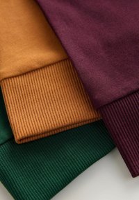Tre ribbede sweatermanchetter i grøn, rustorange og bordeaux, der viser en blød tekstur og en markant farvekombination.