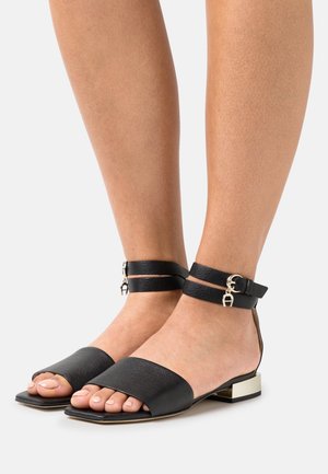 Mustat nahka sandaalit, joissa on leveät etustrapit ja nilkkahihnat. Varustettu kultaisen sävyisillä solkiyksityiskohdilla ja matalalla lohkaremmillä.