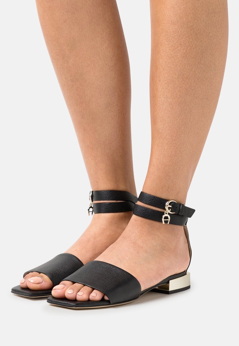 Mustat nahka sandaalit, joissa on leveät etustrapit ja nilkkahihnat. Varustettu kultaisen sävyisillä solkiyksityiskohdilla ja matalalla lohkaremmillä.