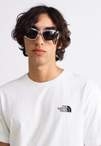 Ochelari de soare negri cu un cadru argintiu, purtați împreună cu un tricou alb simplu, care are un logo negru în zona stângă a pieptului. Textură netedă, design casual.