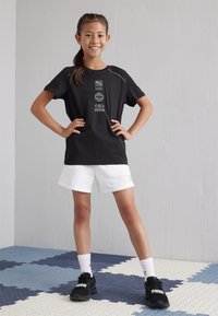 Camiseta negra de manga corta con gráficos del logo gris de Puma, combinada con pantalones cortos blancos y zapatillas deportivas negras con marca visible.