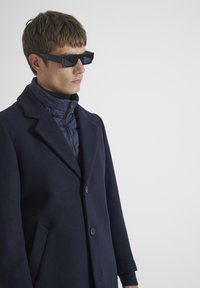Abrigo de lana azul marino con cuello muesca, dos botones y bolsillos laterales sobre una chaqueta acolchada azul marino. El modelo lleva gafas de sol rectangulares negras.