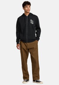 Black zip-up hoodie with a front pocket and white graphic design. Paired with brown trousers and black sneakers. Styled casually.