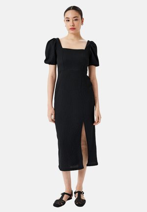 Vestido midi negro con escote cuadrado, mangas abullonadas y abertura lateral. Tejido texturizado y silueta ajustada. Combinado con zapatos negros de tejido trenzado.