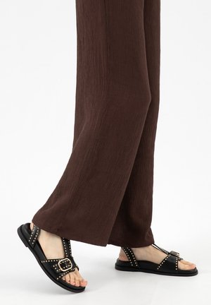 Voeten met zwarte sandalen met studs en gespen, gecombineerd met losse, bruine, gestructureerde broeken tegen een effen witte achtergrond.