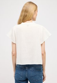Hvid, tekstureret kortærmet top med rund halsudskæring og knapdetalje bagpå, parret med blå jeans med stikninger på baglommerne.
