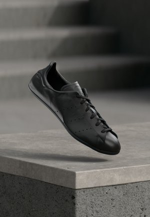 Sneaker in pelle nera con lacci che fluttua sopra una superficie in pietra chiara, con gradini grigi sfocati sullo sfondo.
