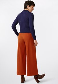 Marineblauer gerippter Pullover mit langen Ärmeln und taillierter Passform; kombiniert mit weit geschnittenen orangen Hosen und dunkelbraunen Blockabsatzstiefeln.
