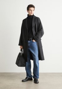 Grey wool overcoat, black turtleneck, blue denim jeans, and black leather shoes. Holds a textured black bag, standing against a neutral backdrop.