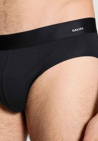 Schwarze Baumwollslips mit einem weichen elastischen Bund, auf dem der Markenname "CALIDA" in Weiß gedruckt ist. Glatte Textur mit minimalen Nähten.