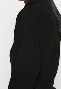 Pull en tricot noir avec un col haut et des épaules tombantes. Le tissu semble doux, texturé et légèrement épais, avec une taille ceinturée.