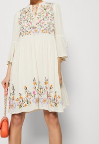 Krämfärgad, flödande klänning med blommig broderi, trekvarts ärmar i klockform och knytkrage. Innehåller en dekorativ tofsdetalj.