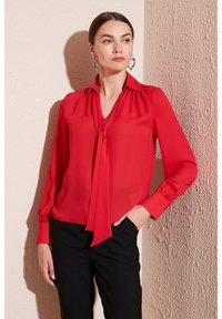 Red blouse made of lightweight fabric with a tie detail at the collar, long sleeves, and gathered shoulders, paired with black trousers.