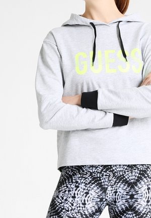 Person trägt einen hellgrauen Hoodie mit "GUESS" in Gelb und schwarz gefärbten Leggings, verschränkte Arme, steht vor einem weißen Hintergrund.
