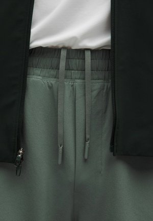 Prim-plan al pantalonilor gri cu elastic la talie și sireturi, asociați cu o cămașă albă și o jachetă neagră cu fermoare.
